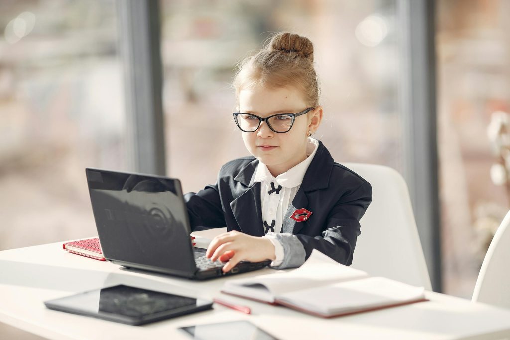 A smartly dressed young girl works on a laptop in a modern office setting.